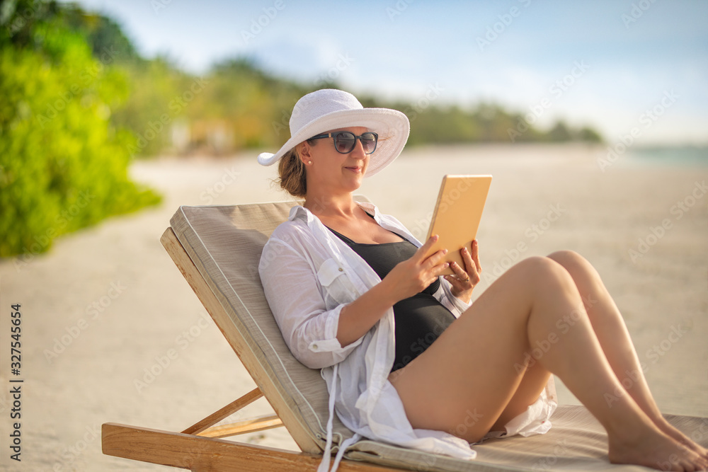 Woman with tablet computer on the beach sitting in the deck chair. Home office concept, passive income concept