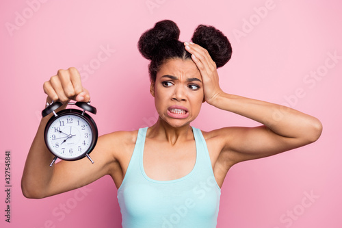 Photo of funny dark skin lady hold metal classic alarm clock hand on head grimacing oversleep missed work afraid to be late wear blue tank-top isolated bright pink background