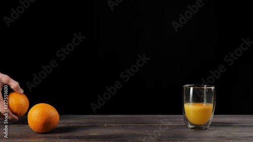 Wide shot of hand takes oranges from the table and throws them in a double-walled glass in turn, after the oranges reach the glass, orange juice appears against black background