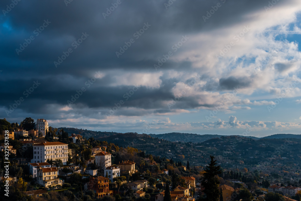 The panoramic cityscape view of a Côte d'Azur town's houses and the Alps mountains next to the Meditarrenean sea under a moody cloudy sky (Grasse, France)