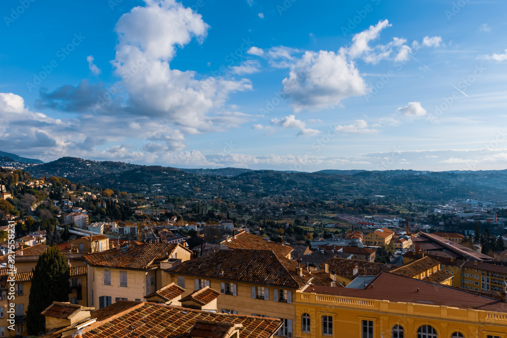 The panoramic cityscape view of a Côte d'Azur town center and the Alps mountains next to the Meditarrenean sea under the blue somewhat cloudy sky (Grasse, France)