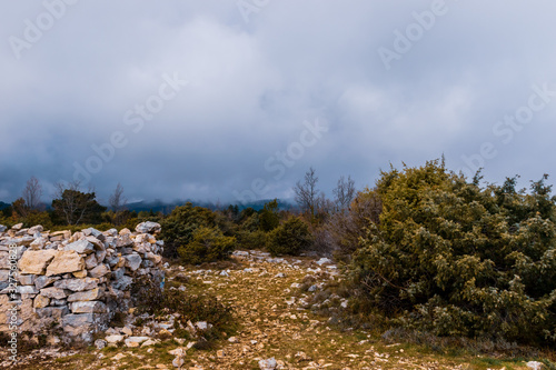 The hiking path on top of the low Alps mountain range under a moody cloudy sky with an old half-ruined stone wall on the left