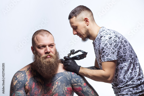 Tattooer artist beats a tattoo to a bearded man in sterile gloves. Tattoomachine. The process is painful. Pain from drawing with a needle. Informal punk rocker. stabbed the chest photo