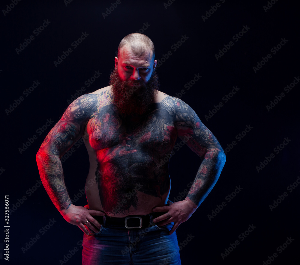 Tattooed brutal bearded man with a naked torso. Tattoo model portrait. Tattooing art artist. Brutal rocker biker stern big dork serious dangerous. Black background. Beard. Tattooed brutal bearded man with a naked torso. Tattoo model portrait. Tattooing art artist. Brutal rocker biker stern big dork serious dangerous. Black background. Beard.