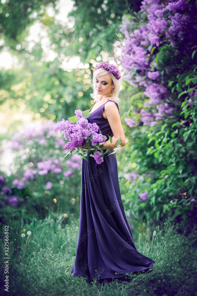 Outdoor fashion photo of a beautiful young woman surrounded by purple flowers. Girls in a long dress with a slit on the background of a spring garden with lilacs. The concept of cosmetics and perfumes