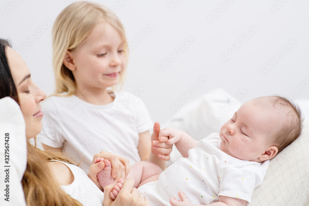 Young mother holding a newborn son in her arms smiling at her elder daughter with white hair sitting next to her Young mother holding a newborn son in her arms smiling at her elder daughter with white hair sitting next to her