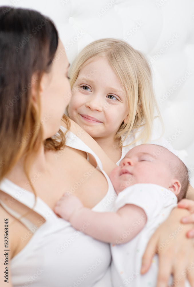 Young mother holding a newborn son in her arms smiling at her elder daughter with white hair sitting next to her
