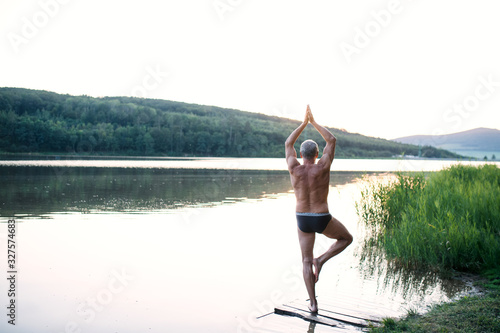 Rear view of senior man in swimsuit standing by lake outdoors doing yoga.