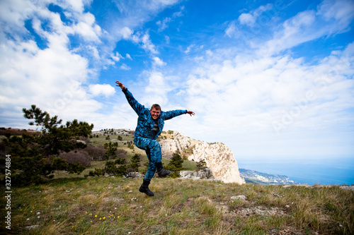 Funny young cheerful caucasian man jumping on the hills with green grass against a blue sky and white clouds. Concept of long-awaited travel and tourism