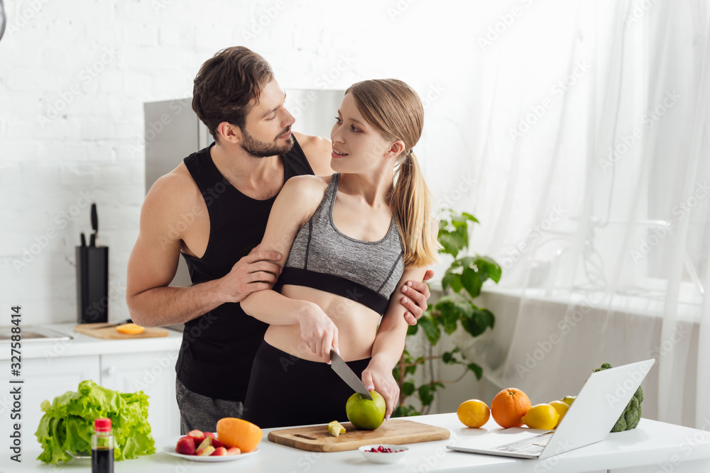 happy girl cutting apple near sportive man, laptop and fruits in kitchen