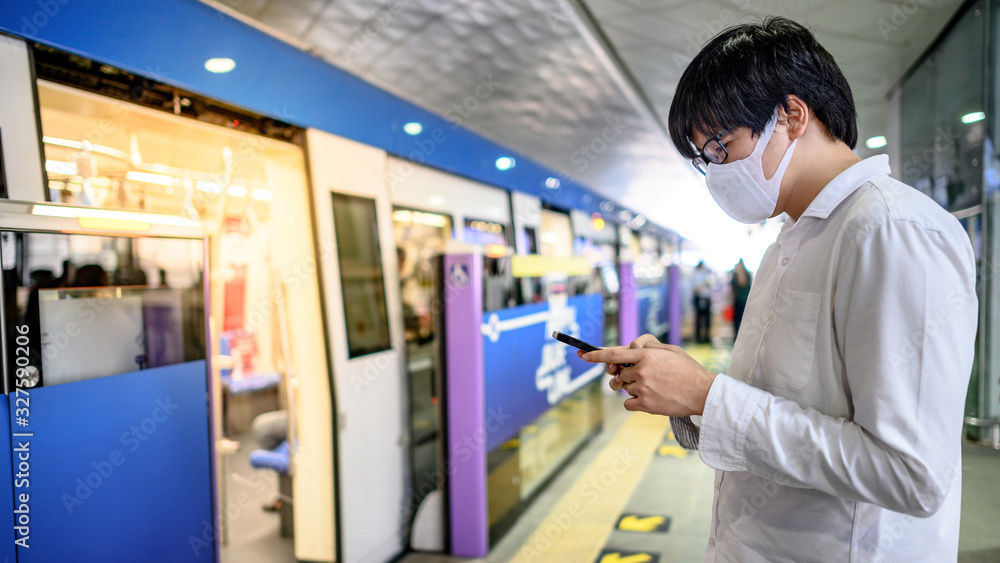 Obraz premium Asian man wearing surgical face mask using smartphone at skytrain station platform. Wuhan coronavirus (COVID-19) outbreak prevention in public transportation. Health awareness for pandemic protection