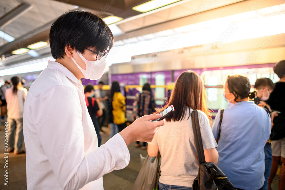 Asian man wearing surgical face mask using smartphone at skytrain station platform. Wuhan coronavirus (COVID-19) outbreak prevention in public transportation. Health awareness for pandemic protection Asian man wearing surgical face mask using smartphone at skytrain station platform. Wuhan coronavirus (COVID-19) outbreak prevention in public transportation. Health awareness for pandemic protection