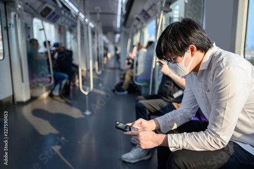 Asian man wearing surgical face mask using smartphone on skytrain or urban train. Wuhan coronavirus (COVID-19) outbreak prevention in public transportation. Health awareness for pandemic protection