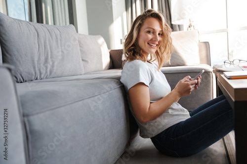 Image of happy woman smiling and using cellphone while sitting on floor