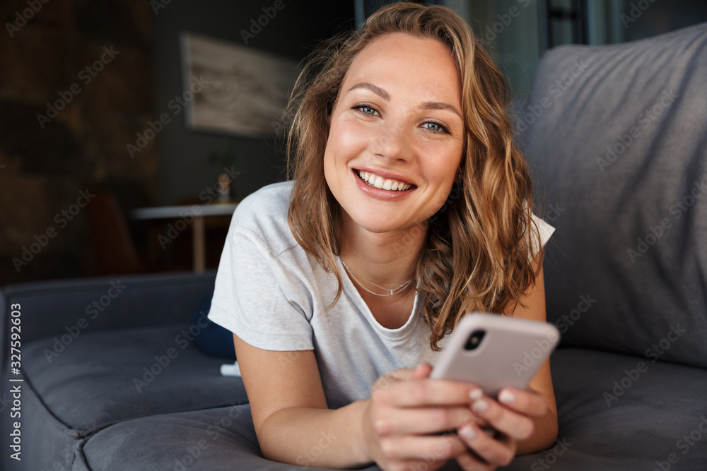 Image of happy woman smiling and using cellphone while lying on sofa - obrazy, fototapety, plakaty 