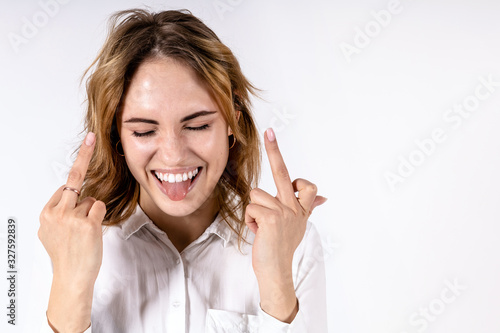 young beautiful girl in a white shirt with a bold smile and sticking out tongue showing fuck off sign to the camera on a white background, copy space