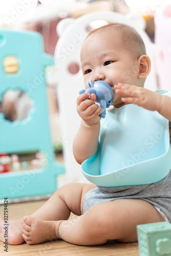 An Asian baby sits in a pen, biting a pacifier