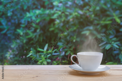 Image of clean white coffee cup or teacup and the plate with soft stream smoke put on old wooden table and blurry fresh green leaves tree in natural background with sunrise lighting and copy space.