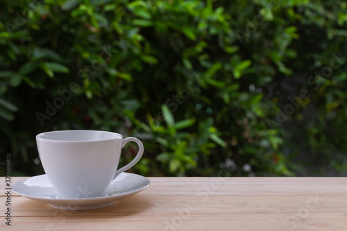 Image of clean empty white coffee cup or teacup and the plate put on old wooden table and blurry dark green leaves tree in natural background with morning soft lighting and copy space.