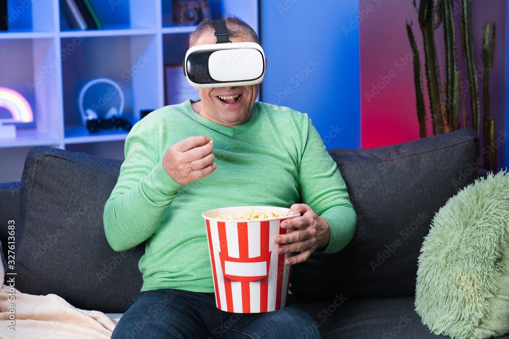Senior Caucasian Man With a Box of Popcorn in His Hand Watching Video Using Virtual Reality Headset Sitting on Sofa Eating Popcorn in Room.