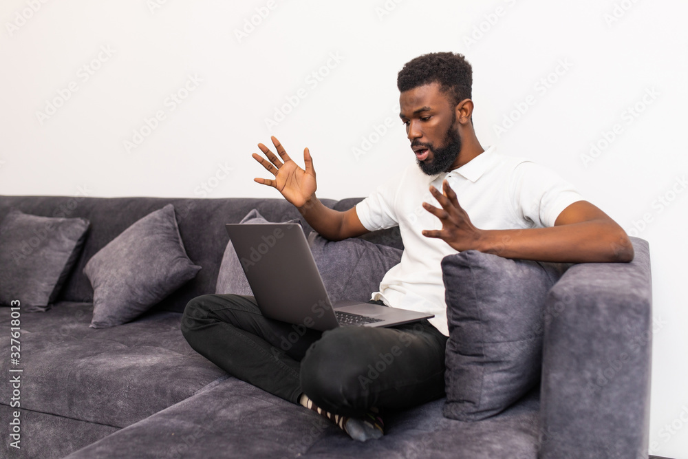 Young shocked black man chatting online on laptop with friend at home. Casual guy sitting on beige couch in light livingroom