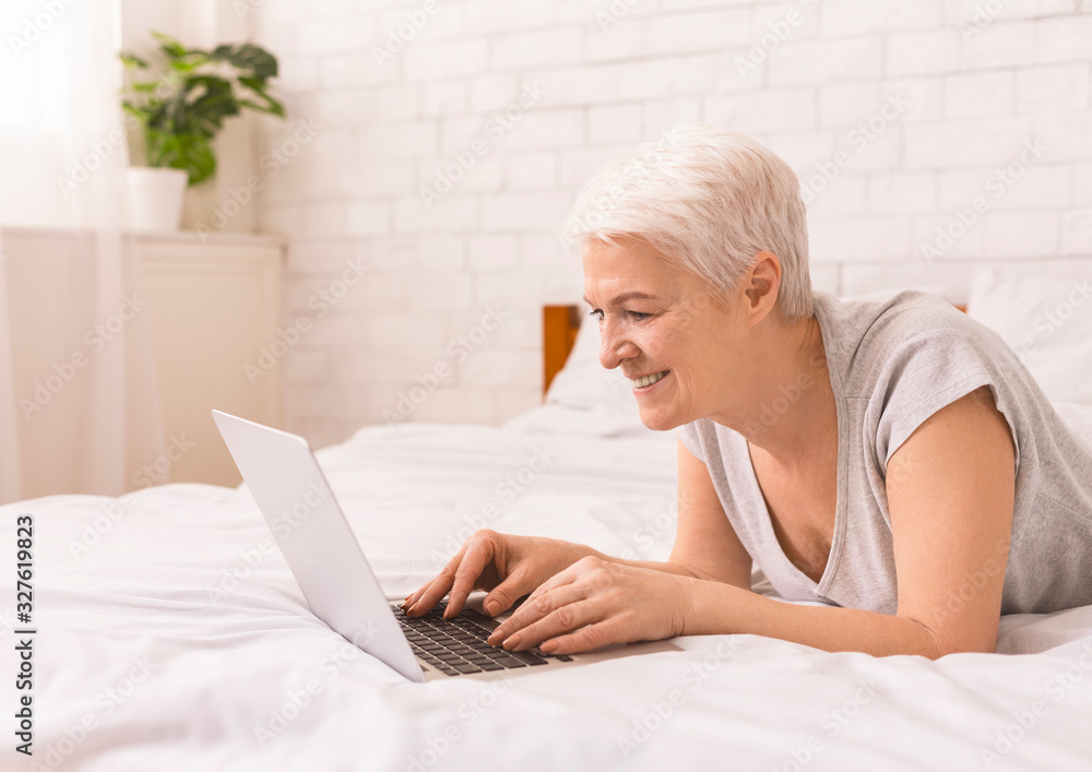 Positive senior woman lying in bed and typing on laptop Positive senior woman lying in bed and typing on laptop