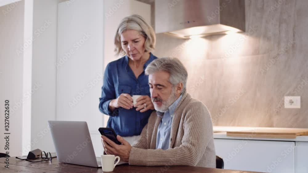 Mature couple using laptop and smartphone at table in the kitchen Mature couple using laptop and smartphone at table in the kitchen