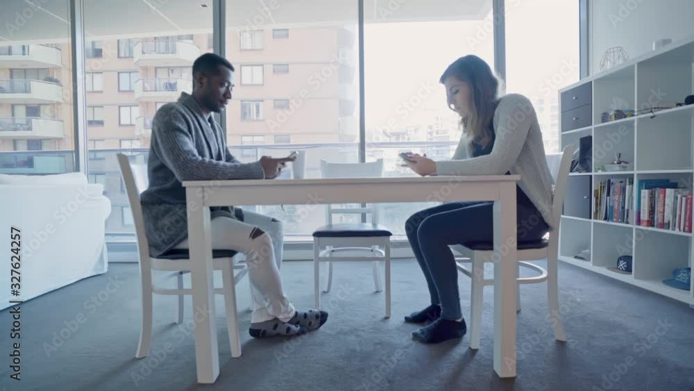 Millennial multiracial couple with a smartphone addiction sitting at a kitchen table, ignoring each other. Millennial multiracial couple with a smartphone addiction sitting at a kitchen table, ignoring each other.