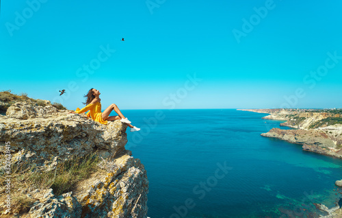 side view of traveler woman in bright yellow dress sitting on cliff edge with beautiful sea view and enjoying wonderful nature