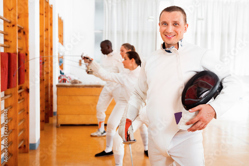 Active young male fencer in uniform standing with mask and foil at fencing room