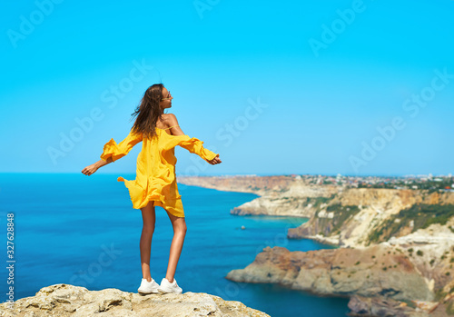 Full length portrait of taned traveler woman standing on top rock beach with open arms, bright yellow dress and hair blowing in the wind.