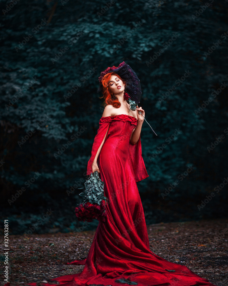 Amazing beautiful cute young woman with fiery red hair in red long dress stands on path in green forest with bouquet of red roses in her hands. Amazing beautiful cute young woman with fiery red hair in red long dress stands on path in green forest with bouquet of red roses in her hands.