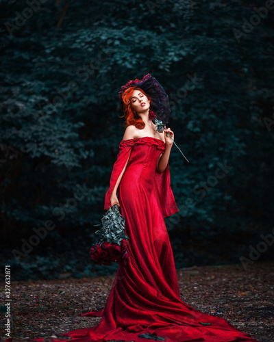 Amazing beautiful cute young woman with fiery red hair in red long dress stands on path in green forest with bouquet of red roses in her hands.
