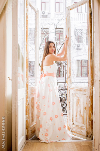 Young affectionate woman in flower dress standing on balcony at home. Beautiful fashionable girl in stylish spring dress posing at vintage studio. Pretty fashion model posing in vintage interior © Artem