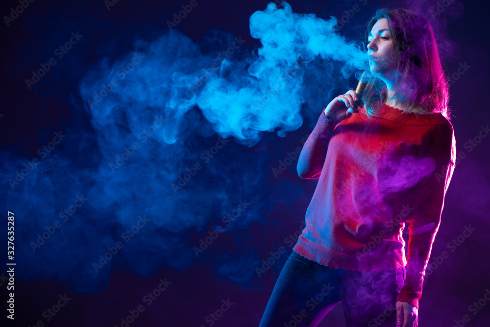 Woman viper on a dark background. Vaper in the clouds of steam. Steam next to a smoking woman. Concept - modern cigarettes. Concept - giving up cigarettes in favor of vape. Young girl with an e-cig