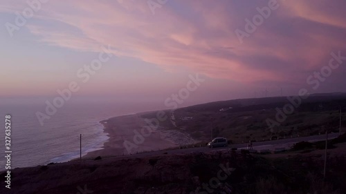 Panoramic aerial shot of car driving down the street in front of a cliff with the coastline and a colourful sunset in the backdrop. photo