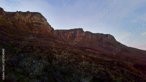 Table Mountain and Oudekraal Nature reserve during golden hour. Aerial drone pan shot revealing the silhouette of the rocky brown coastal mountains against blue sky during sunset. photo