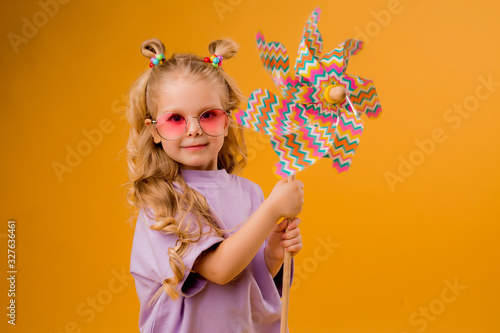 little blonde girl smiling in sunglasses holding a windmill stands on a yellow background in the Studio isolate. happy child, text space, horizontal photo