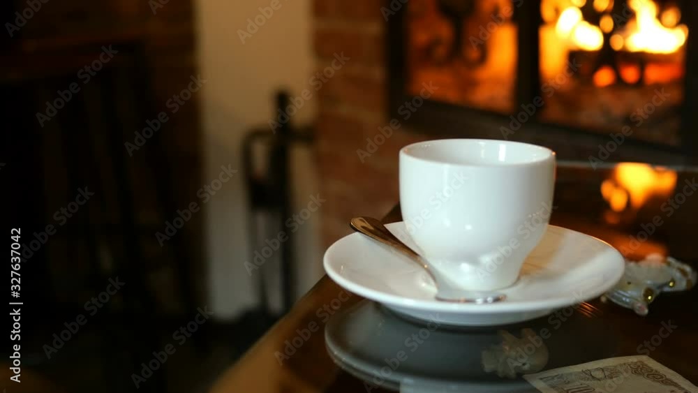 Cup of coffee is on table in cafe or restaurant with fireplace. Glimmers of flame reflect on the table. Romantic setting. Selective focus. Close-up. Cup of coffee is on table in cafe or restaurant with fireplace. Glimmers of flame reflect on the table. Romantic setting. Selective focus. Close-up.