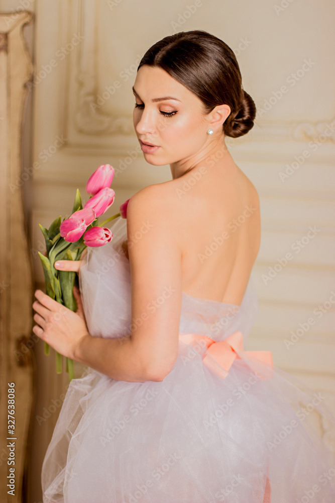 Portrait of affectionate and beautiful girl in white dress holding tulips in hands and posing with closed eyes indoors. Stylish pretty woman with spring bouquet of flowers. Young bride. 8 march Portrait of affectionate and beautiful girl in white dress holding tulips in hands and posing with closed eyes indoors. Stylish pretty woman with spring bouquet of flowers. Young bride. 8 march