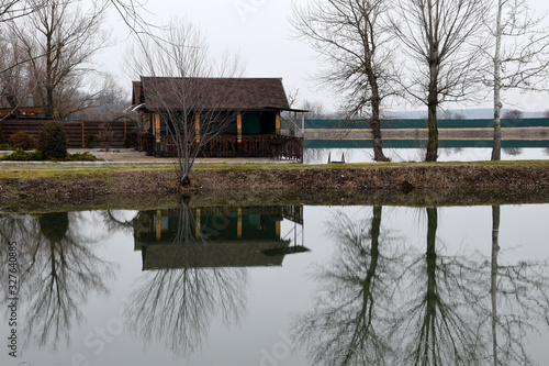 A wooden gazebo stands on the lake. Trees and bushes are reflected in the water surface. On the street is early spring or autumn.
