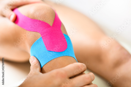 Kinesiology taping treatment with blue and pink tape on athlete patient injured knee. Woman hands apply kinesio treatment after sports muscle injury. photo