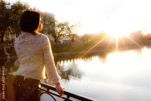 Side view of a young dreamy brunette girl standing on a bridge and enjoying the sunset by the river on a sunny summer evening. Concept of expectation of change and new © YouraPechkin