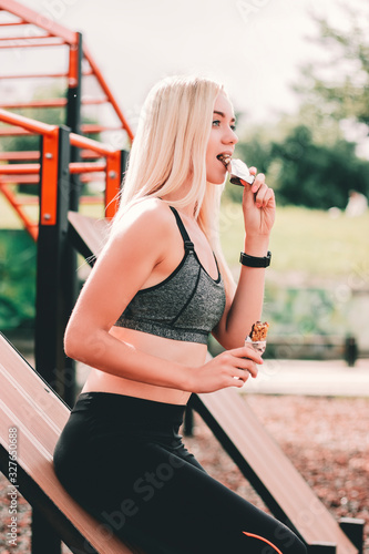 Attractive young hungry fitness woman enjoying healthy muesli or cereal snacks, having rest after workout training outdoors. Beautiful blonde sports girl posing and eating protein bar on sports ground