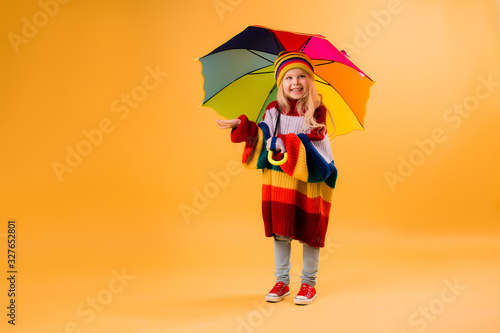 cute little girl in a knitted sweater and hat is holding a multicolored umbrella on a yellow background