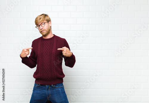 young blonde man looking proud, positive and casual pointing to chest with both hands agaist vintage tiles wall photo