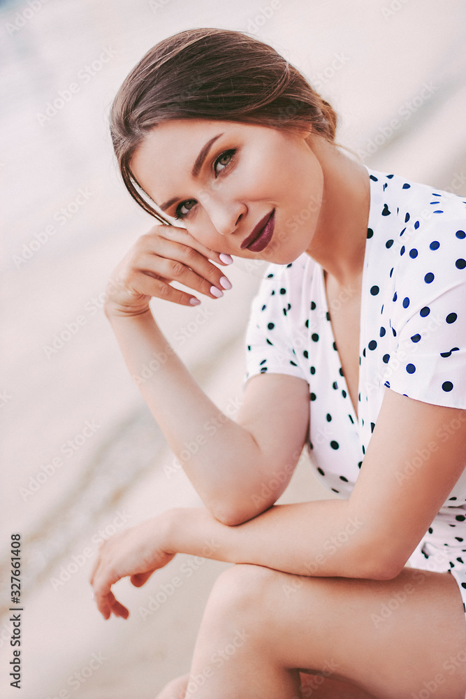 Portrait beautiful gentle girl in white polka dot dress sitting on sandy beach and smiling during evening romantic after party outdoors. Young tender woman in stylish retro clothing posing at seaside Portrait beautiful gentle girl in white polka dot dress sitting on sandy beach and smiling during evening romantic after party outdoors. Young tender woman in stylish retro clothing posing at seaside