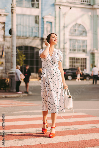Young attractive woman retro model in white polka dot dress, red high heels crossing street with handbag in hand. Beautiful affectionate girl in retro clothing posing, move on city road. Noise effect