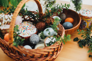 Traditional Easter basket. Easter modern eggs, easter bread, ham, beets, butter, in wicker basket decorated with green boxwood branches and flowers on rustic wooden table