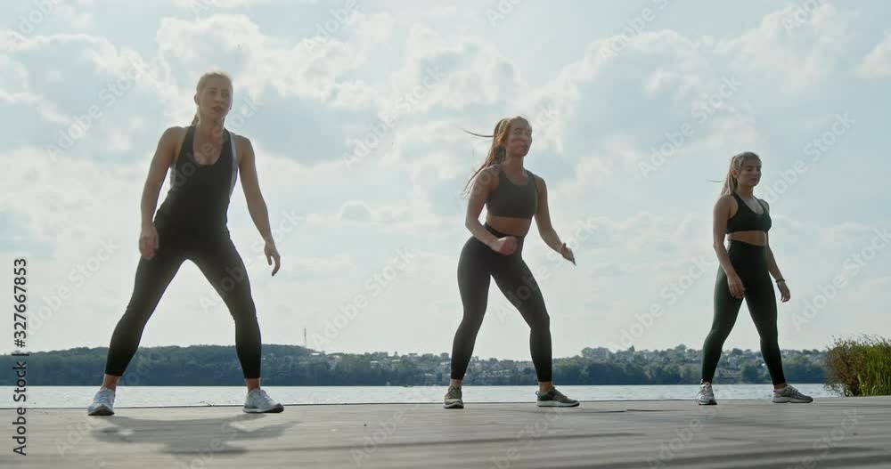 Low angle of young slim women in sports outfit training together and performing squat exercise at riverbank in sunny summer day Low angle of young slim women in sports outfit training together and performing squat exercise at riverbank in sunny summer day